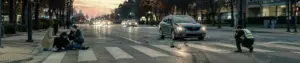 Silver sedan on a crosswalk after a collision as two bystanders comfort an injured person sitting on the curb, while a photographer in a reflective vest crouches to take photos at dusk.